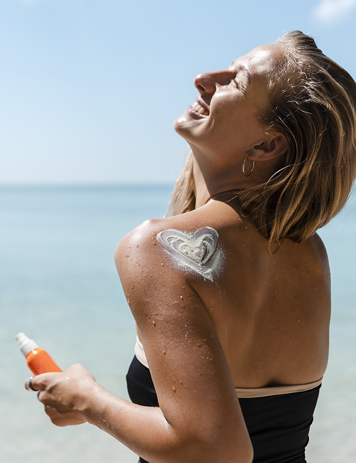 Woman smiling at the beach with a bottle of sunscreen and cream on her back.